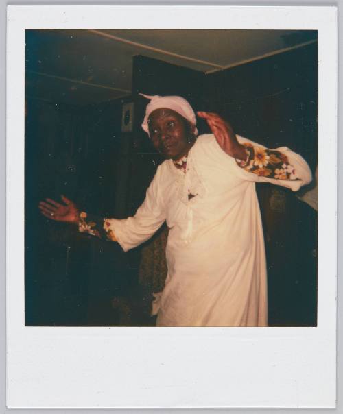 [Woman in white blouse with headscarf in dark room with arms raised to her shoulders]