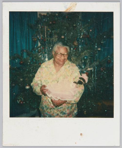 [Elderly woman smiling in her pajamas, standing in front of Christmas tree with doll in her arms]