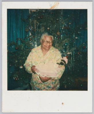 [Elderly woman smiling in her pajamas, standing in front of Christmas tree with doll in her arms]