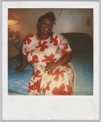 [Woman sitting on bed in red and white floral dress holding a pink rose]