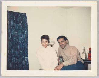 [Woman and man in turtleneck sweaters sit next to each other, man is holding a blue glass with ice inside of it]