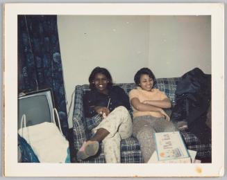 [Two young women smiling and sitting on couch together, a board game inside of its' box sits in front of them]