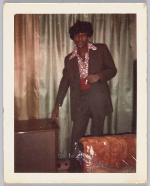 [Man in hat having a cigarette in living room, two fingers placed on table beside him]