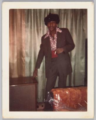 [Man in hat having a cigarette in living room, two fingers placed on table beside him]