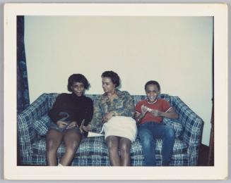 [Two women and a boy sitting on couch together, woman in middle has a magazine open and is smiling at woman on the left]
