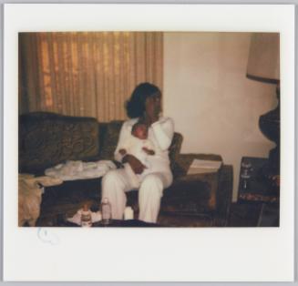 [Woman in white sweatsuit sitting on couch with baby on lap, brushing hair out of her face]