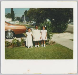 [Three young girls dressed in white standing in row on lawn in front of sidewalk, brown car in background]
