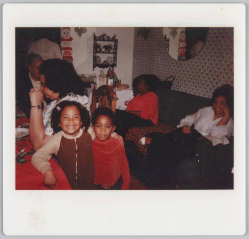 [Young girl and boy smiling with arms around each other, women sitting on couch in background]