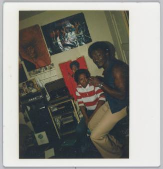 [Man and woman sitting on right side of room, posters are hung on wall behind, record player in shelf to the left of them]
