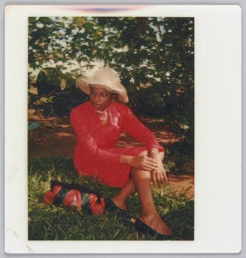 [Woman wearing red dress and yellow hat sitting and holding her knees on grass]
