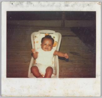 [Baby in white onesie sitting in car seat with hands in fists outstretched]
