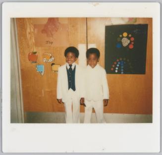 [Two boys standing together wearing white suits in front of wall with children's art posted on it]
