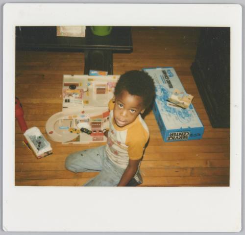 [Boy sitting on floor looking up at photographer, a toy set of a train is beside him]
