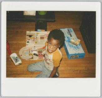 [Boy sitting on floor looking up at photographer, a toy set of a train is beside him]
