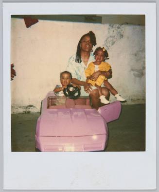 [Woman sitting on back of toy car with two toddlers, one sitting on her lap, the other inside the car]
