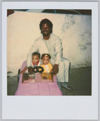 [Man sitting on back of toy car with two toddlers sitting inside]
