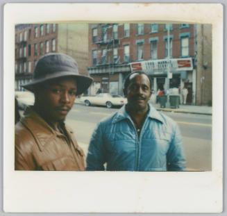 [Two men dressed in jackets standing on city street, food market in background]

