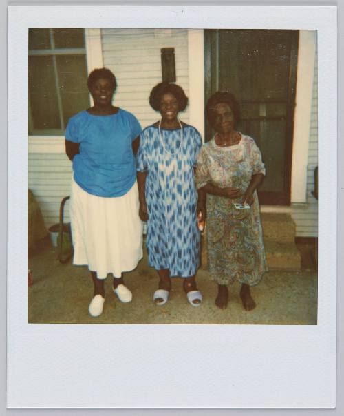 [Three middle-aged women standing in a row outside of home with door and window behind them]
