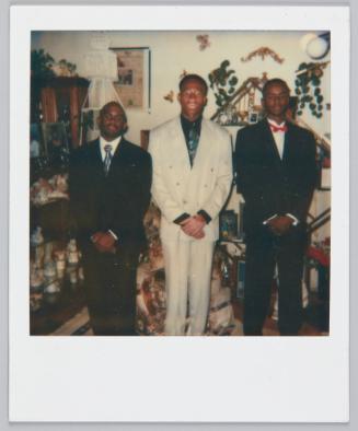 [Three men standing in room with many knick-knacks, all three men have their hands clasped in front of them, wearing suits]
