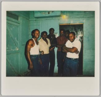 [Group of men standing together inside, man on left in white tank top and man on right has his arms crossed]