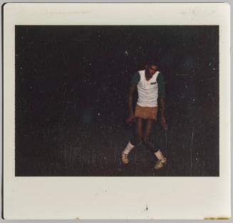 [Adult in white top, shorts, and white socks and shoes standing with knees together in a dark space]