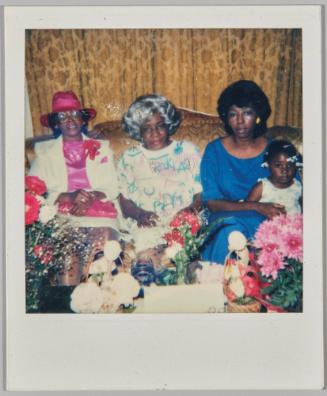 [Three women sitting on couch, woman on left in pink hat and woman on right in blue dress holding toddler, table with flowers in front]