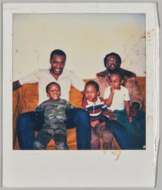 [Two men and three boys on orange couch, boy in centre in red, blue and white striped collared shirt and making funny face]