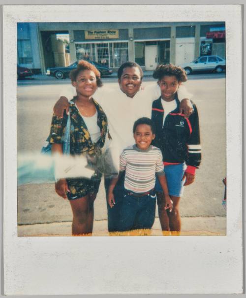 [Man with his arm around a woman to the left and a teenage boy to the right, boy in striped t-shirt standing in front, they are on the sidewalk]