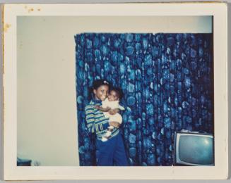 [Young woman in striped shirt holding a baby and standing in front of a blue floral curtain]