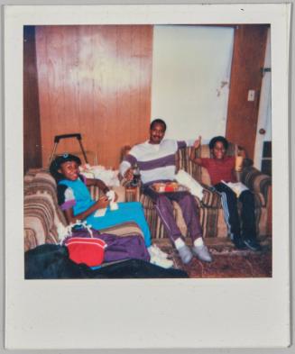 [Man, girl and boy sitting on matching striped couches in wood paneled room, the man has a tray on his lap]