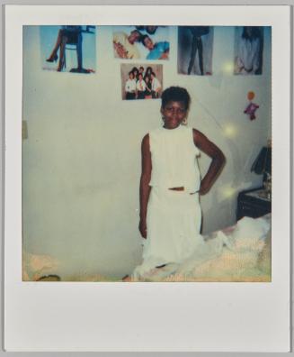 [Girl in white outfit standing with her hand on hip, posters on the wall behind her]