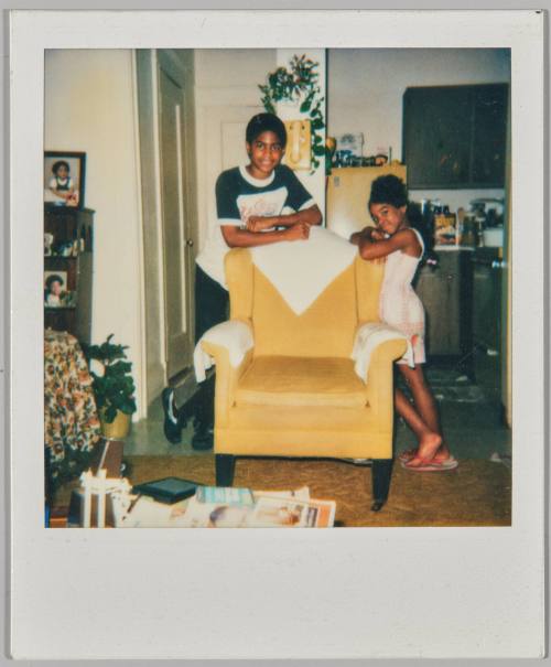 [Boy and girl leaning on a yellow armchair with white coverings, yellow phone on the wall behind them]