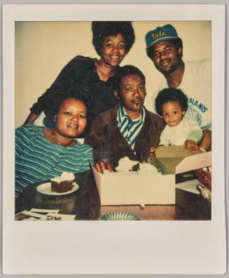 [Four adults and a baby posing around a table with a box of cake, man standing on the right in "Ucla" baseball cap]