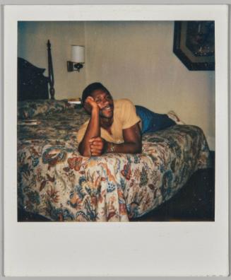 [Man in yellow t-shirt lying on top of bed with his head in his hand]