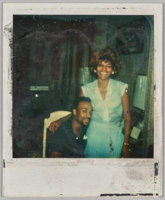 [Woman in blue dress standing with her arm around a man sitting down]