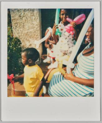 [Woman and children outside with animal balloons, boy in yellow t-shirt holding a red balloon and other child holding a green and pink balloon]