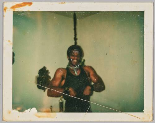 [Man in black tank top with his hands in fists, he is in the corner of a room and a rope is hanging behind him]