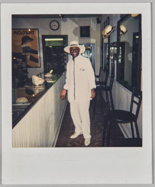 [Man in white brimmed hat, zip-up jacket and pants standing by a bar]