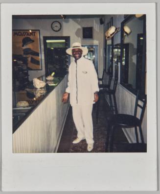 [Man in white brimmed hat, zip-up jacket and pants standing by a bar]