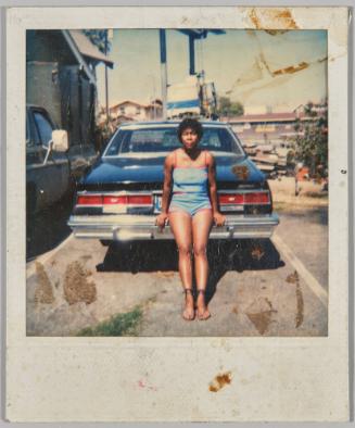[Woman with short hair in blue and red tank top and shorts leaning against the trunk of a parked car]