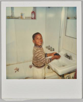 [Boy smiling at camera, he is at the bathroom sink with the tap on]