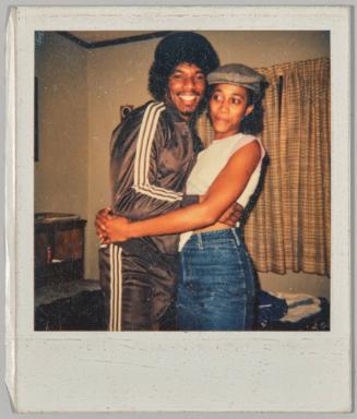 [Man in striped tracksuit hugging woman in grey cap]