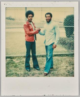 [Two men in front of chainlink fence shaking hands, man on the left in flared denim jeans]