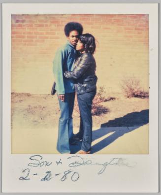 Son + Daughter 2-2-80 [Man in denim jacket hugged by woman in black jacket outside in front of brick wall]