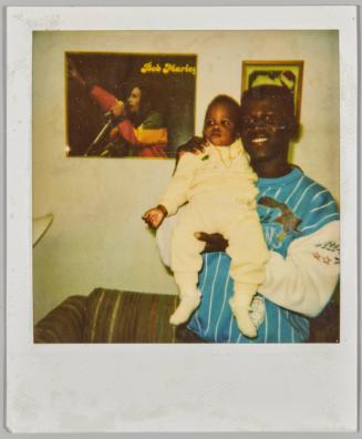 [Man in blue and white sweatshirt holding up a baby with a gold necklace]