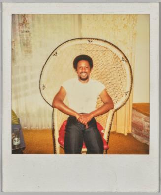 [Man in white t-shirt and black pants sitting on rattan chair with red cushion]