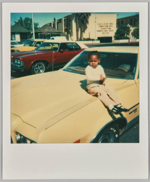 [Child in a white holding their hands together and sitting on the hood of car in parking lot]