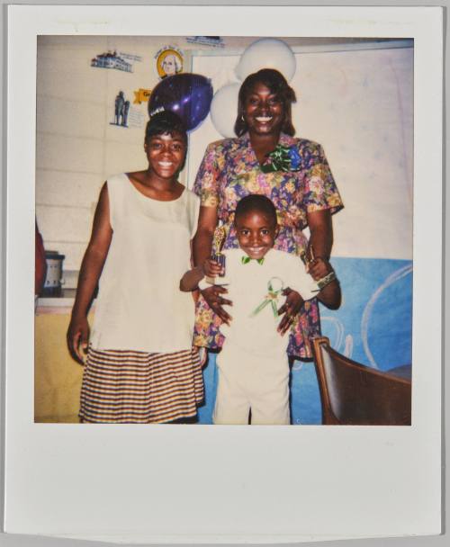 [Two women with a boy wearing a green ribbon, they are standing inside in front of white and purple balloons]