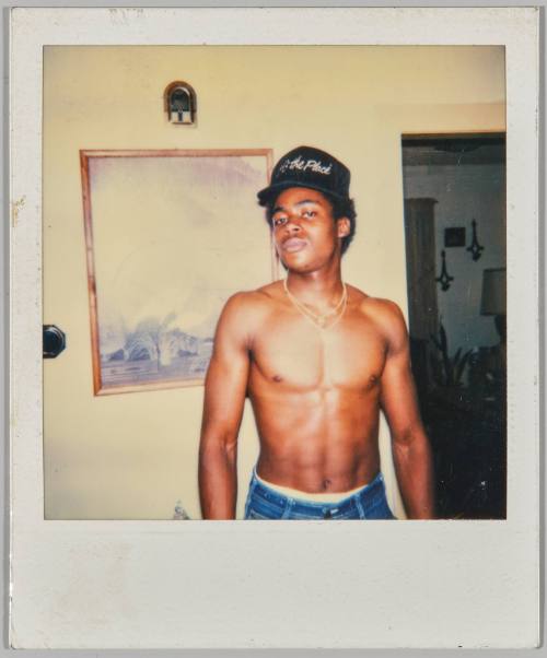 [Shirtless man with black baseball cap and two gold chains standing inside]