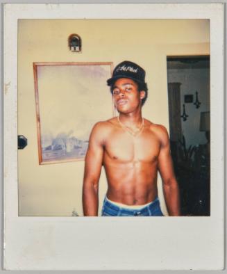 [Shirtless man with black baseball cap and two gold chains standing inside]